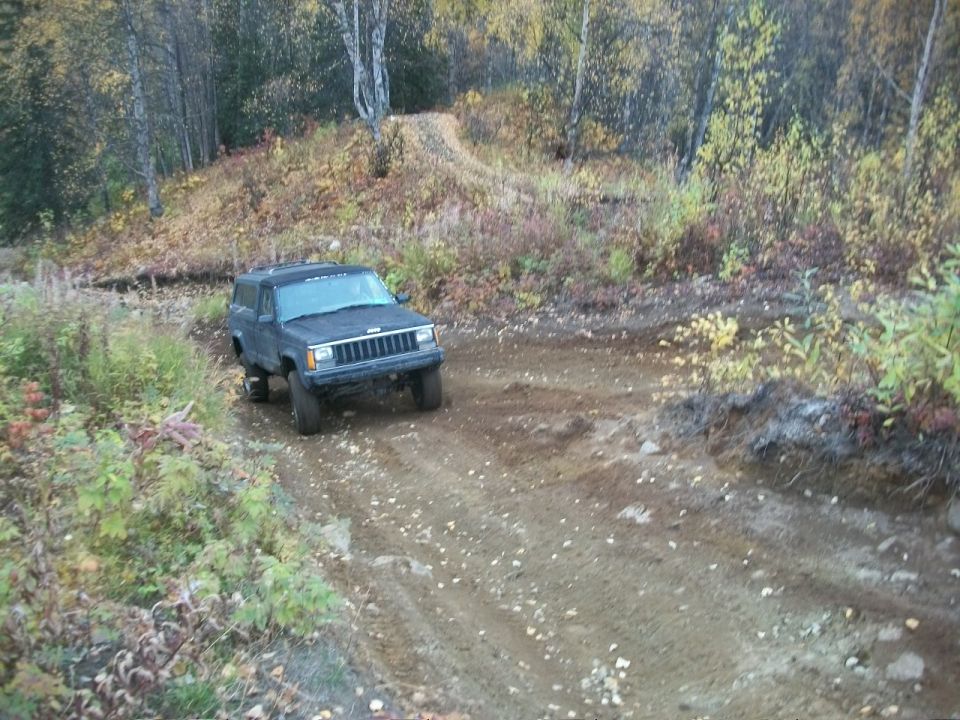 Dark gray Jeep Cherokee XJ on a dirt road, front-left view, with trees and foliage in the background