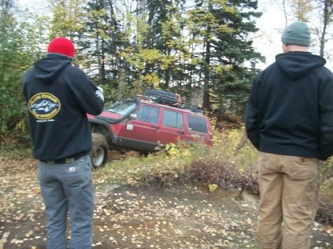 1979 Ford Bronco - Two people watch a red SUV navigate a wooded off-road trail.