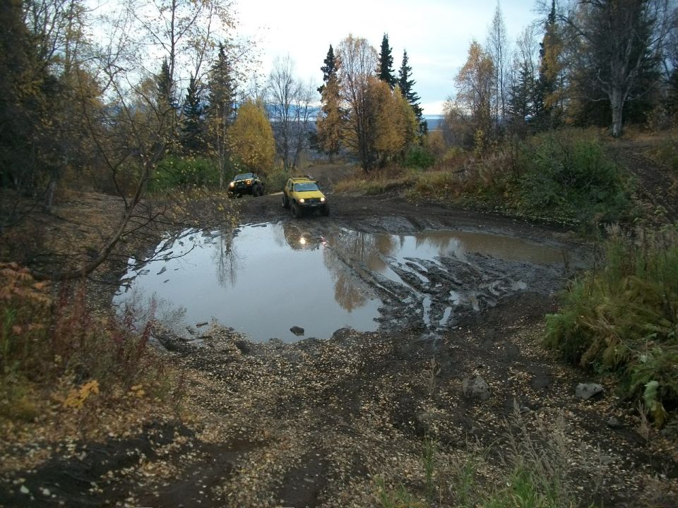Two SUVs navigate a large muddy puddle on a dirt track surrounded by autumn trees