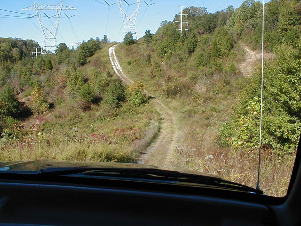 View from inside a vehicle looking out at a winding dirt road going up a grassy hill with power line towers in the background
