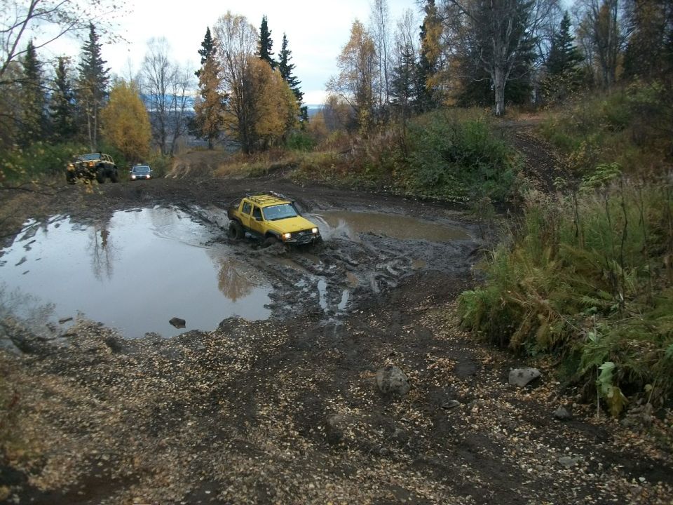 Yellow SUV navigating a muddy puddle on a dirt track, with trees and other vehicles in the background