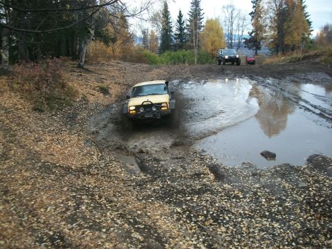 1979 Ford Bronco - A yellow 1978 Ford Bronco navigates a muddy track, kicking up water, with other vehicles visible further down the path.