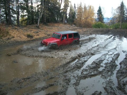 1979 Ford Bronco - A red Jeep Wrangler navigates a challenging muddy terrain, with its front-right wheel deep in the mire.