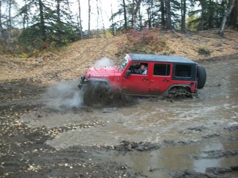 1979 Ford Bronco - A red Jeep Wrangler Rubicon navigates a muddy off-road trail, kicking up water and mud.
