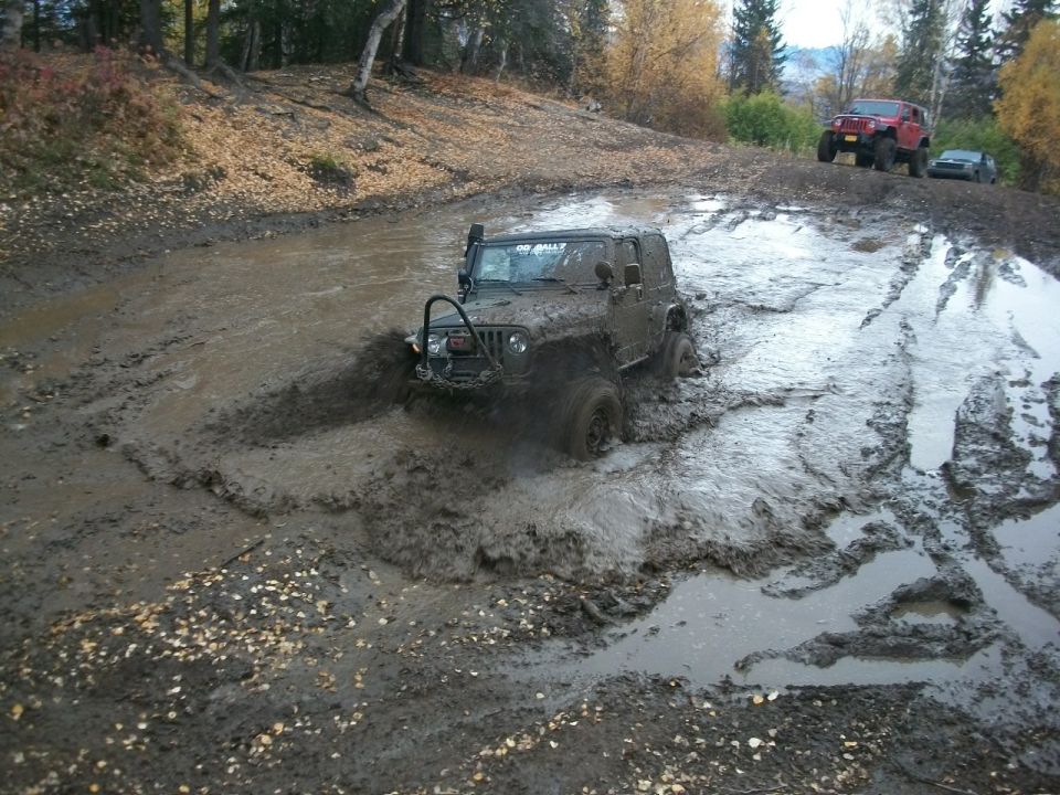 Mud-covered Jeep Wrangler splashing through a muddy track, with a red Jeep and a dark car in the background