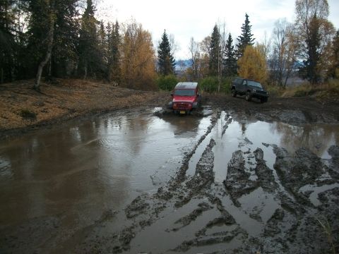 1979 Ford Bronco - Two 4x4 vehicles are shown traversing a challenging muddy terrain with deep tire tracks and standing water.