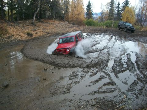 1979 Ford Bronco - A red Jeep Wrangler struggles through a muddy off-road track, with another SUV visible in the background.