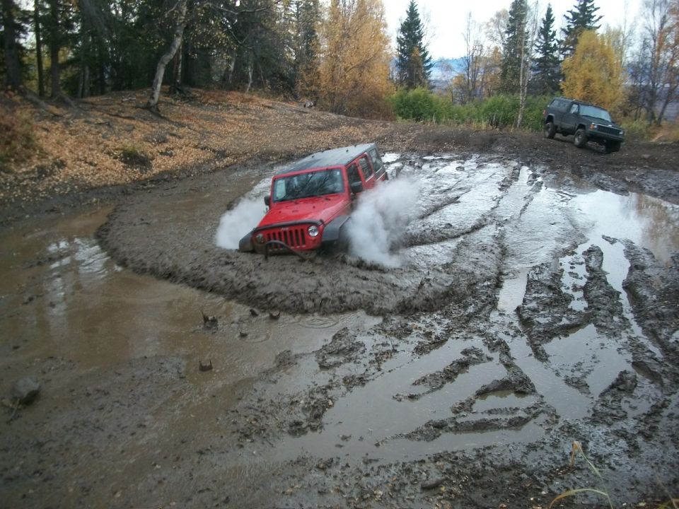 Red Jeep Wrangler with front tires submerged in mud, splashing water