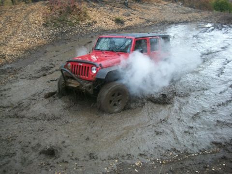 1979 Ford Bronco - A red Jeep Wrangler navigates a muddy trail, creating a cloud of smoke as it churns through the mire.