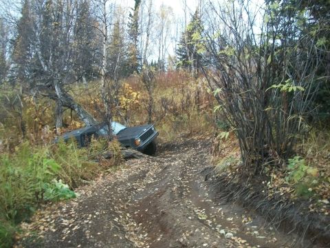 1979 Ford Bronco - An off-road vehicle is stuck on a muddy path, leaning against a tree in a wooded area.