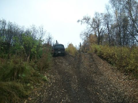 1979 Ford Bronco - A dark SUV navigates an uphill dirt path covered in fallen leaves and mud.