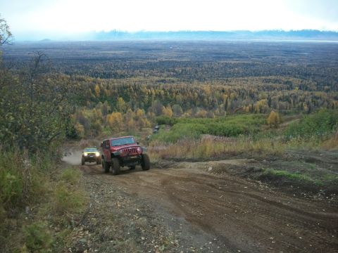 1979 Ford Bronco - Two off-road vehicles navigate a dusty trail through a wooded landscape during autumn.