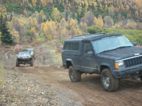 1979 Ford Bronco - Several off-road vehicles navigate a dusty trail through a forest with fall foliage.