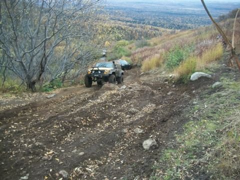 1979 Ford Bronco - Two off-road vehicles traverse a challenging, muddy trail on a steep, wooded slope with autumn foliage visible.