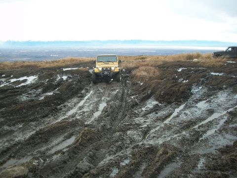 1979 Ford Bronco - A yellow Jeep Wrangler navigates a challenging muddy terrain with tracks from previous vehicles visible.