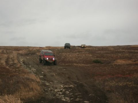 1979 Ford Bronco - Three off-road vehicles traverse a muddy, grassy terrain under an overcast sky.