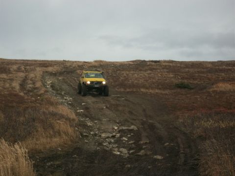 1979 Ford Bronco - A yellow off-road SUV navigates a challenging dirt incline under a cloudy sky.