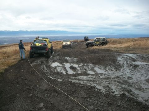 1979 Ford Bronco - Multiple off-road vehicles traverse a challenging muddy trail with a scenic backdrop of water and mountains.