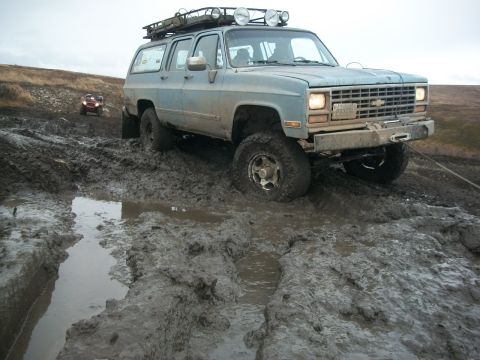 1979 Ford Bronco - This light blue SUV is stuck deep in mud on an off-road trail, with another vehicle visible in the distance.