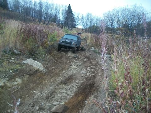 1979 Ford Bronco - Black Jeep Cherokee XJ navigates a challenging, rocky off-road trail with dry vegetation surrounding it.