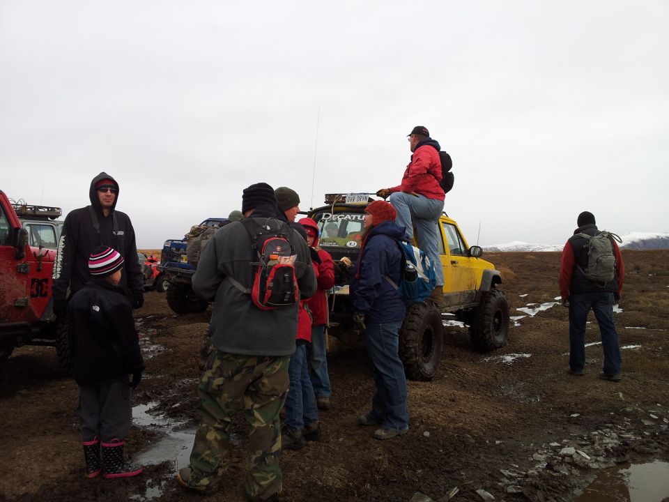 Group of people gathered around several off-road vehicles in a muddy, open field under an overcast sky. One person sits on top of a yellow Jeep