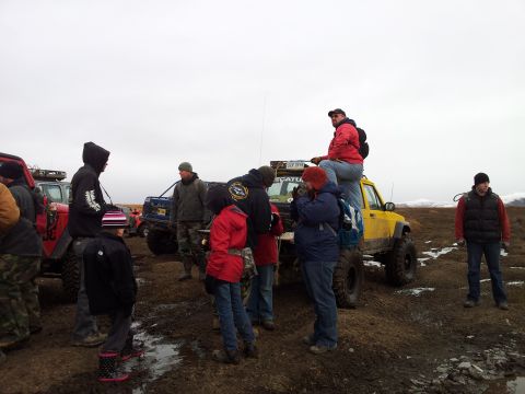 1990 Chevrolet Suburban - A group gathers around several off-road vehicles in a wet, muddy terrain under an overcast sky.
