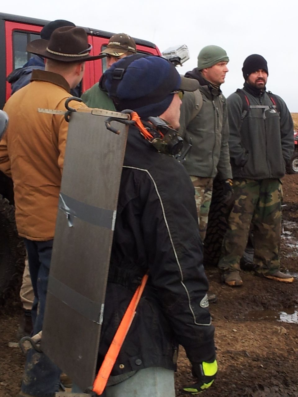 People gathered around a red vehicle and other off-road vehicles in a muddy field