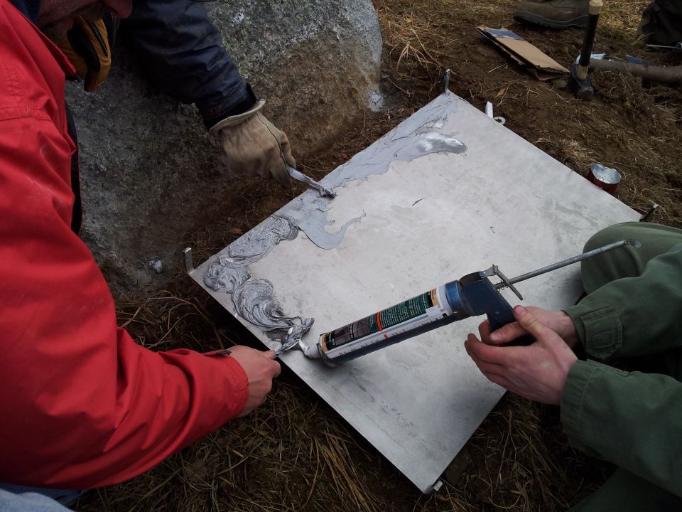 People applying sealant to a square metal plate outdoors