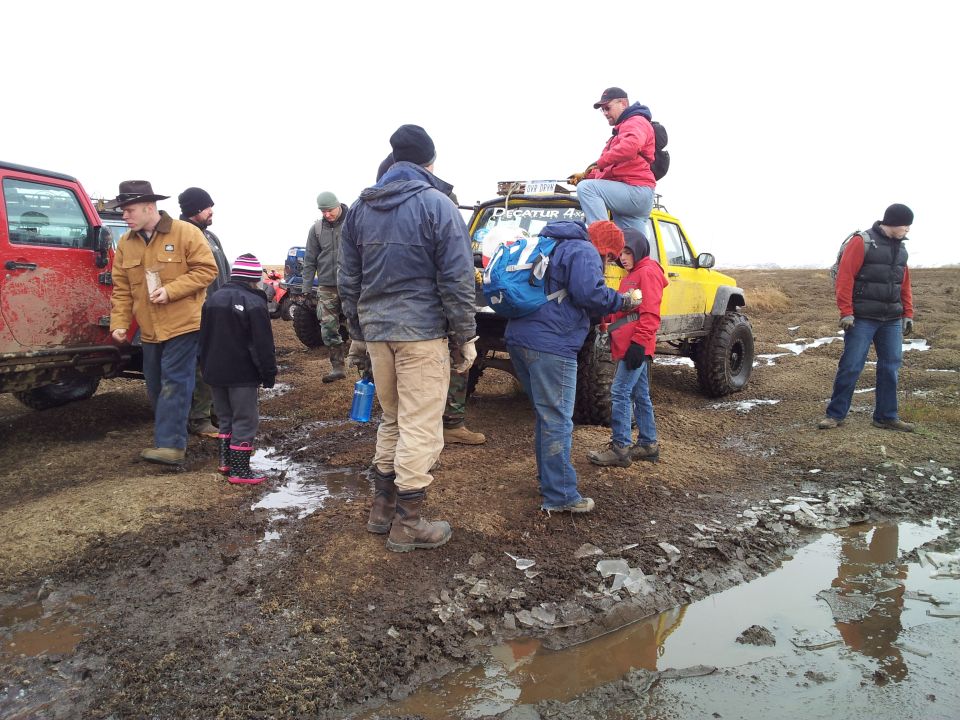 Group of people and off-road vehicles gathered in a muddy, icy area. One person sits on top of a yellow Jeep