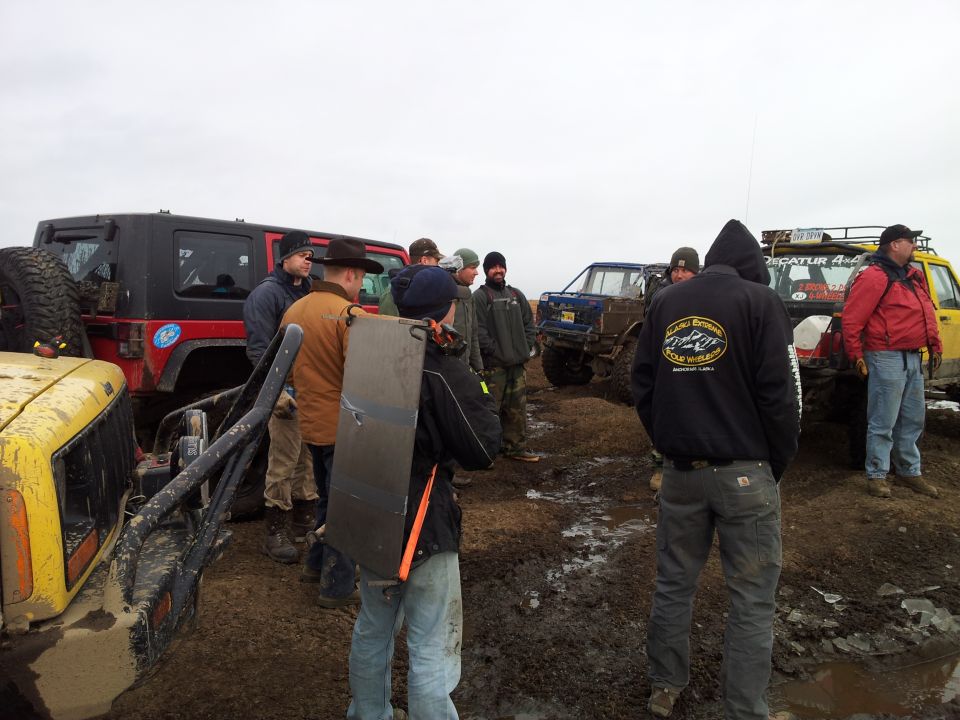 Group of people gathered around several off-road vehicles in a muddy, outdoor area