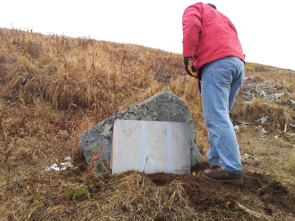 Person in red jacket and jeans digging next to a large rock with a taped-on sign