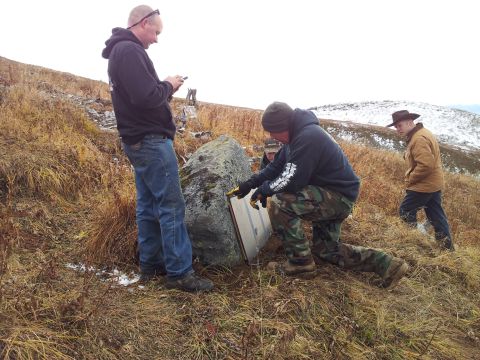 1990 Chevrolet Suburban - Men investigate a metal plate attached to a rock on a hillside with sparse vegetation and distant snow-capped mountains.