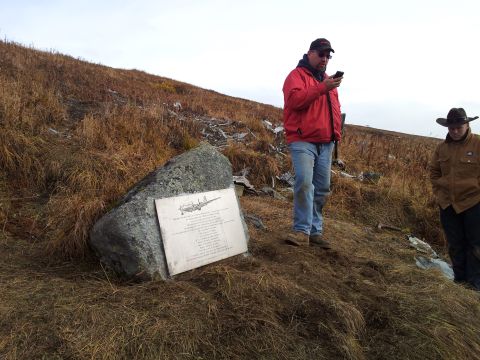 1990 Chevrolet Suburban - Memorial plaque with airplane drawing and text on a rock, two men standing nearby on a grassy hillside.