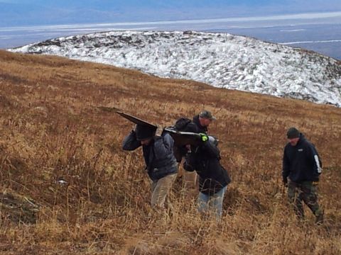 1990 Chevrolet Suburban - People navigate a grassy, snow-dusted hillside carrying a large object.