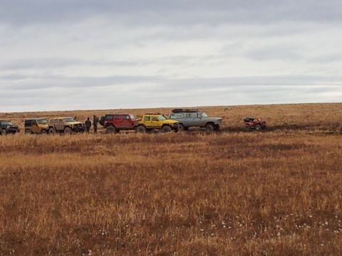 1990 Chevrolet Suburban - Several off-road vehicles, including a gray SUV, are lined up in a desolate, grassy landscape under an overcast sky.
