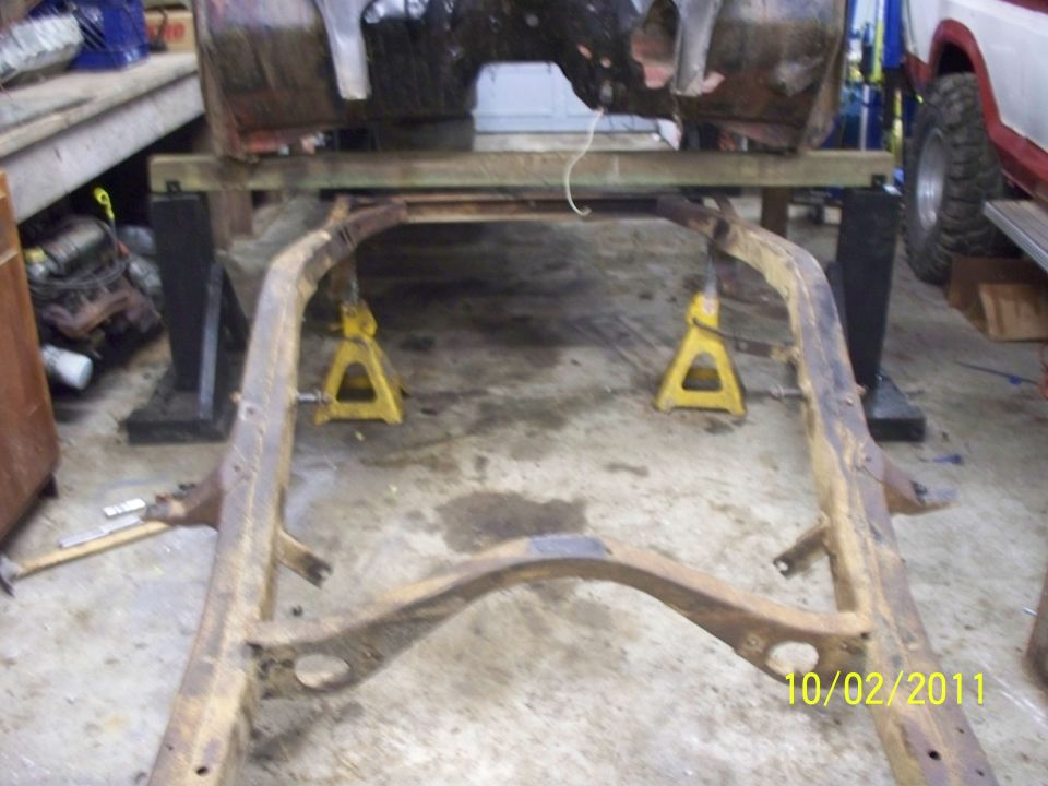Vehicle frame supported by jack stands in a garage. A partial vehicle body is visible above the frame