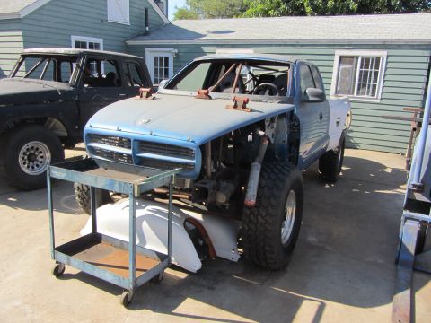 1987 Suzuki Samurai - A partially disassembled blue pickup truck with a roll cage and large off-road tires sits in a yard next to another vehicle.