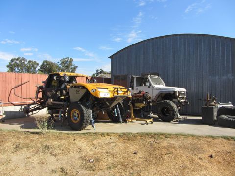 1987 Suzuki Samurai - Two off-road vehicles, one a buggy and the other a white SUV, are parked outside a large metal building.