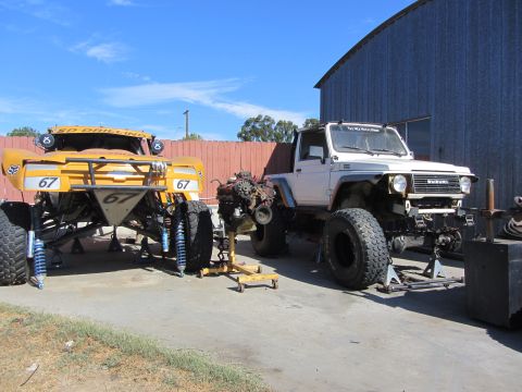 1987 Suzuki Samurai - Two off-road vehicles, one yellow buggy and a white Suzuki Samurai, are parked outside a metal building.
