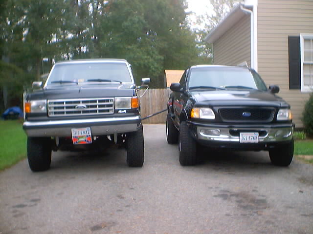 Black lifted Ford Bronco parked beside a black Ford F-150 pickup in a driveway, front view