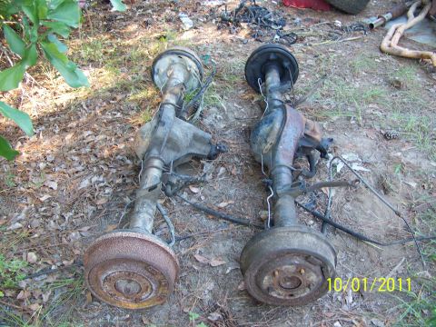 1986 Ford Bronco - Two rusty rear axle assemblies with drum brakes are shown lying on the ground outdoors.
