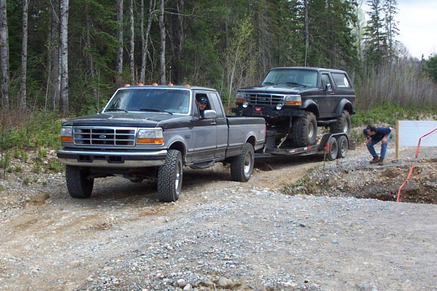 Gray Ford F-Series 4x4 pickup towing a lifted Bronco on a trailer down a gravel road