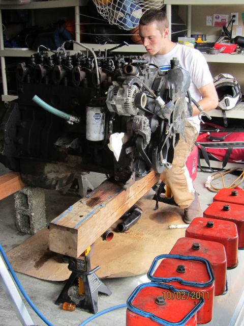 1984 Ford Bronco - An engine is supported by jack stands as a person works on it, with red fuel cans lined up nearby.