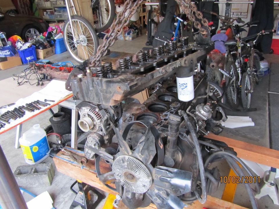 Engine block with valve covers removed, suspended by chains, with fan and alternator visible