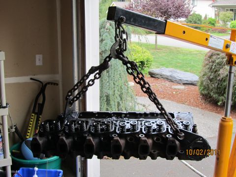 1984 Ford Bronco - Engine cylinder head suspended by chains from a yellow engine hoist, with a garage interior and outdoor greenery in the background.