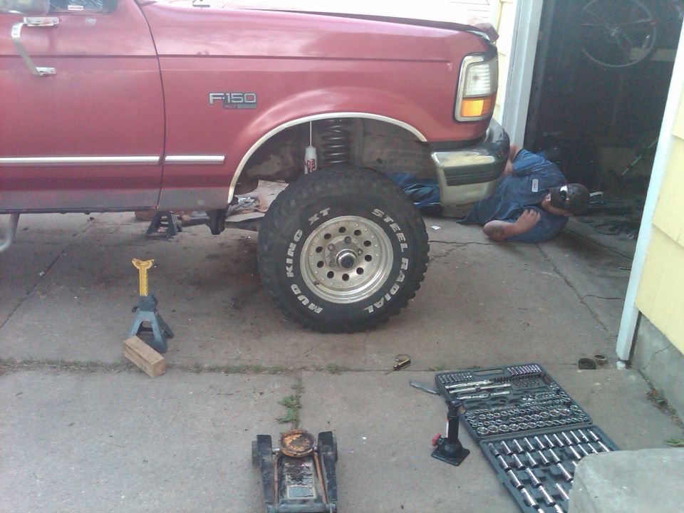 Person under red Ford F-150 on jack stands with tools and removed wheel