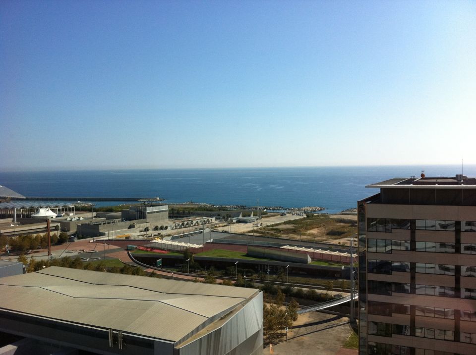View of a coastal city with buildings and the ocean under a clear blue sky. Watermark reads SUPERMOTORS