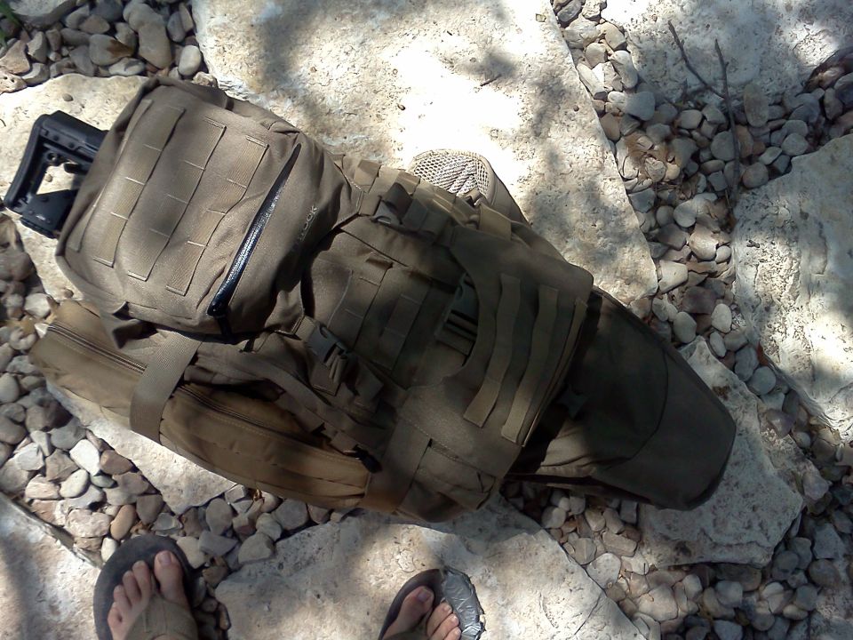 Olive drab tactical backpack with MOLLE webbing and a rifle stock protruding from the top, resting on rocks