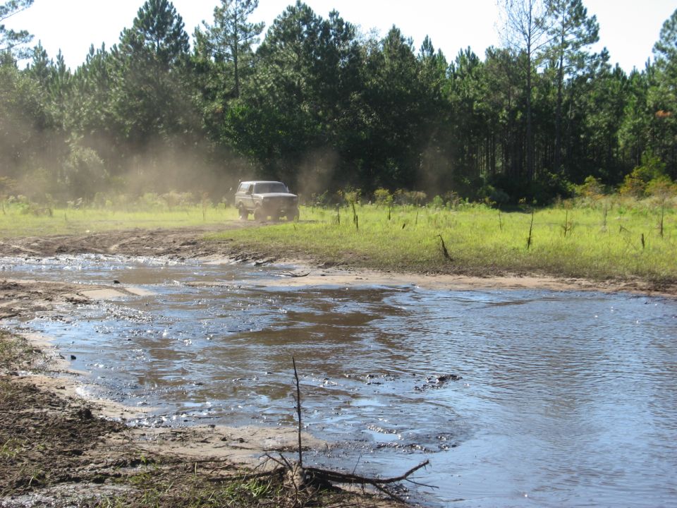 1994 Ford Bronco kicking up dust driving through a muddy field near trees