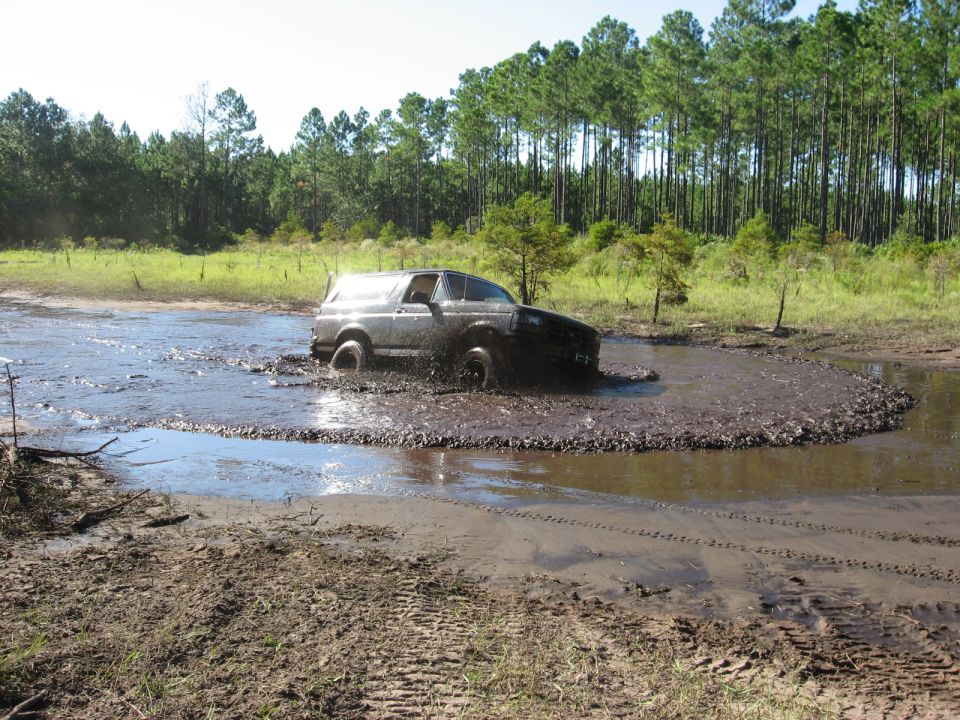 1994 Ford Bronco driving through a muddy puddle with trees in the background